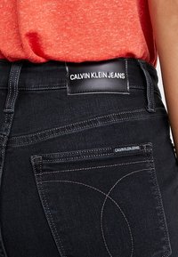 Vaqueros de denim negro con una etiqueta de cuero en la parte trasera que dice "CALVIN KLEIN JEANS". Presentan costuras naranjas y un diseño ajustado.