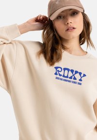 Beige Sweatshirt mit lockerer Passform, blauem "ROXY"-Logo und dem Text "SURFING COMPANY SINCE 1990". weicher Stoff und gerippter Kragen. Baumwollmischung.