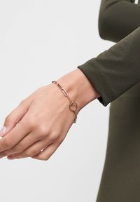 Guldtonet metalarmbånd med aflange papirklem links og en cirkulær accent, båret på et håndled med en olivengrøn langærmet top.
