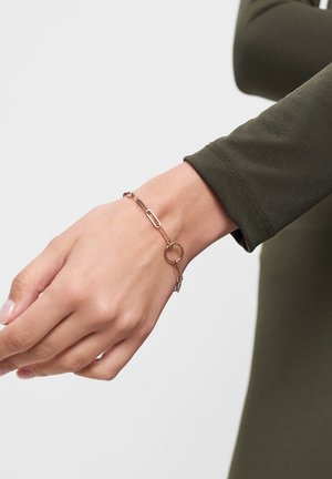Armband aus goldfarbenem Metall mit länglichen Paperclip-Gliedern und einem runden Akzent, getragen an einem Handgelenk mit einem olivgrünen Langarmoberteil.