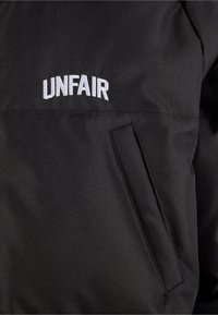 Svart stoffjakke med teksturert finish, med brodert "UNFAIR"-logo i hvitt. Inkluderer en skjult sid lomme.
