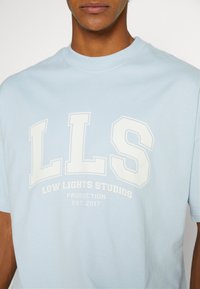 Personne portant un T-shirt bleu clair avec le texte blanc "LLS Low Lights Studios Production Est. 2017" sur la poitrine.