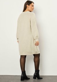 Abito maglione beige con accenti a coste su spalle e polsini. Maniche lunghe e taglio alla lunghezza del ginocchio. Indossato con stivali neri alla caviglia.