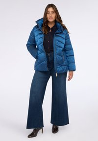 Chaqueta de plumas azul con alto cuello, cierre de cremallera y patrón de acolchado horizontal. Combinada con pantalones de mezclilla acampanados de tiro alto y botas de tacón.
