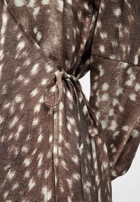 Robe portefeuille en tissu marron avec un motif à pois blancs, nouée à la taille par une fine ceinture en tissu formant un nœud en forme de boucle.