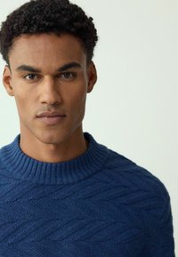 Granatowy sweter dziany z prążkowanym, okrągłym dekoltem. Z przodu ma fakturowany wzór jodełki. Miękki materiał.