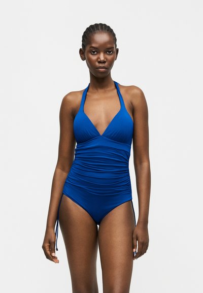 Femme aux cheveux tressés portant un maillot de bain une pièce bleu à encolure américaine froncée, debout devant un fond clair uni.