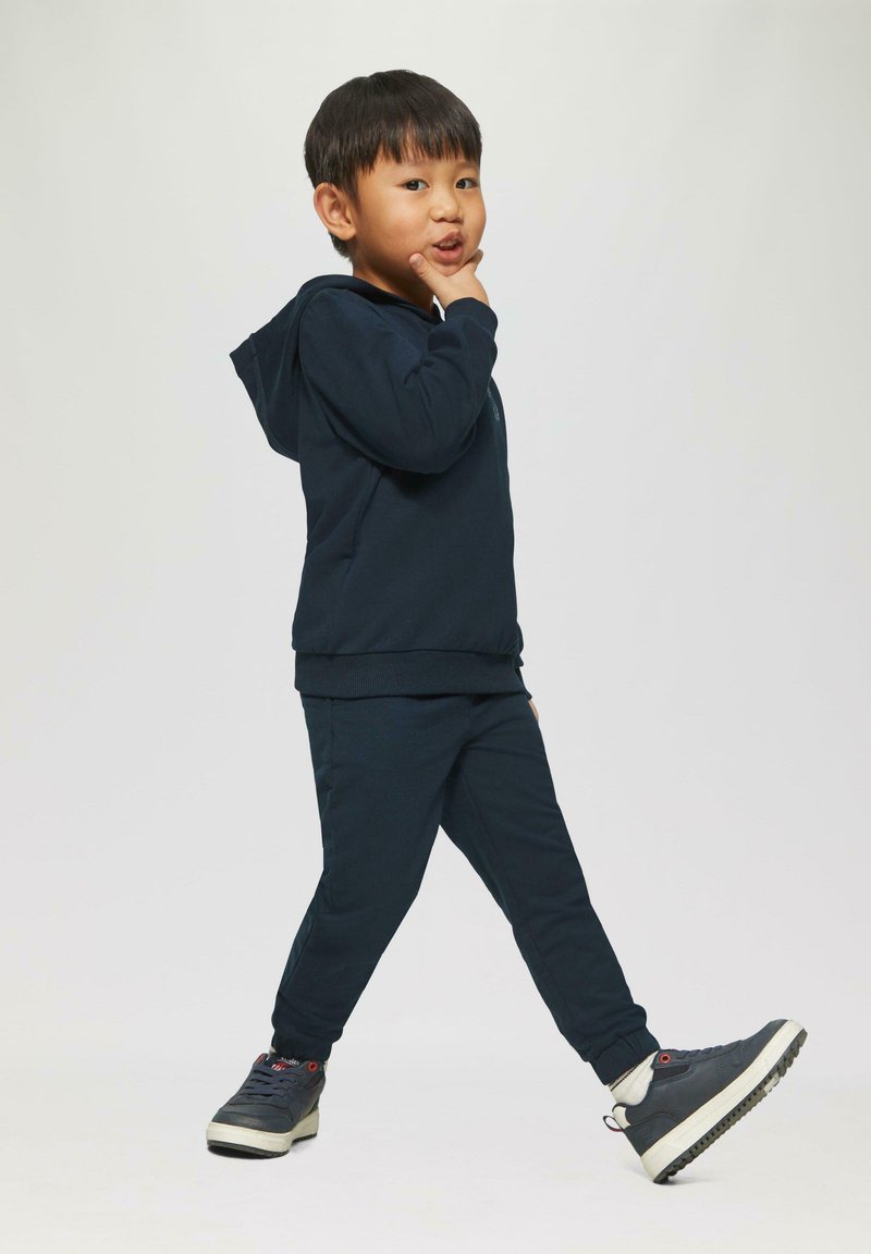 Jongen in een marineblauwe hoodie en broek, die met één been naar voren loopt, met zijn hand op zijn kin rustend, en iets opzij kijkt.