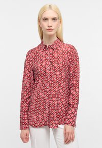 Rotes Langarmhemd mit einem geometrischen weißen Muster, mit Kragen und Knopfleiste. Kombiniert mit weißen Hosen.