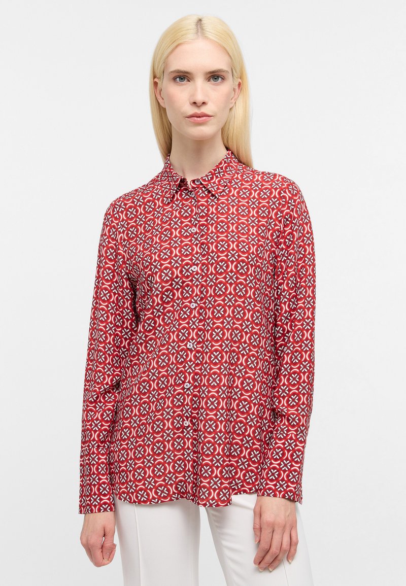 Rotes Langarmhemd mit einem geometrischen weißen Muster, mit Kragen und Knopfleiste. Kombiniert mit weißen Hosen.