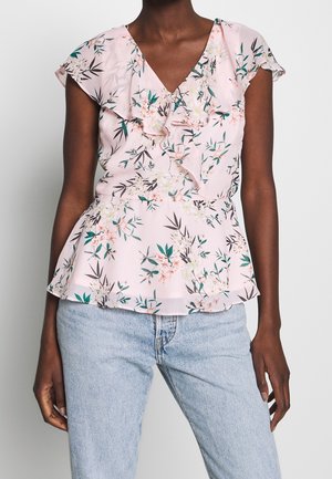 Femme portant un chemisier floral rose clair avec un col en V à volants et des manches courtes, associé à un jean bleu clair sur un fond uni.