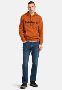 Pomarańczowy hoodie z niebieskim logo, przednia kieszeń i sznurowany kaptur. Połączony z niebieskimi dżinsami oraz czarnym i brązowym obuwiem.