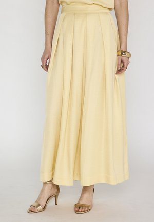 Donna con gonna plissettata a vita alta color giallo pallido abbinata a sandali dorati open-toe e braccialetto dorato al polso destro.