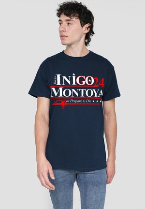 Young man with dark curly hair wears a navy blue T-shirt reading “Vote for Inigo Montoya 24 or Prepare to Die” in red and white text.