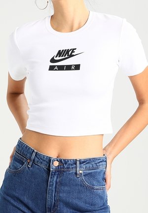 Femme portant un t-shirt court blanc Nike Air et un jean bleu taille haute, mains posées sur les hanches devant un fond uni.