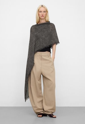 Vrouw met lang blond haar, gekleed in een zwart topje, beige wijde broek, bruine open-toe schoenen en een lange donkere sjaal met franjes over één schouder gedrapeerd.