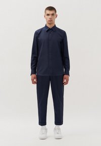 Camicia button-up blu navy con colletto, abbinata a pantaloni tapered coordinati. Sneakers bianche casual completano l'outfit. Tessuto di una consistenza liscia.