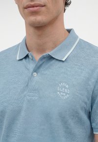 Bărbat purtând un tricou polo texturat de culoare albastru deschis, cu tiv alb la guler și logo "Denim Blend" pe piept, stând în fața unui fundal alb.