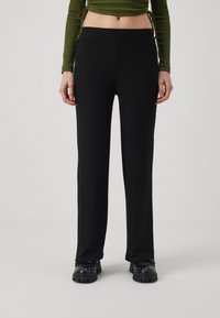 Black ribbed wide-leg trousers with an elastic waistband, featuring a smooth texture. Worn with a green long-sleeve top and chunky black shoes.