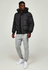 Schwarze Pufferjacke mit gestepptem Muster, Kapuze, Fronttaschen und einem logoed Innenfutter. Kombiniert mit grauen Sweatpants und weißen Turnschuhen.