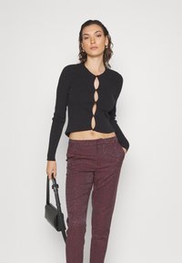 Μαύρο κοντό πουλόβερ με στρογγυλή λαιμόκοψη και πέντε οβάλ ανοίγματα ασφαλισμένα με κουμπιά. Συμπληρωμένο με μπορντώ μοτίβο παντελόνι και μαύρη τσάντα.