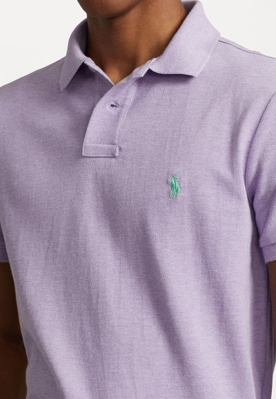 Lilla polo särk, mis on valmistatud tekstureeritud kangast, kahe nööbiga kraega ja vasakul rinnal rohelise tikandiga logoga.