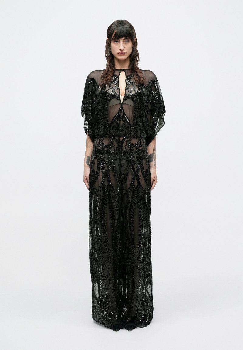 Sötét hajú nő áll fehér háttér előtt, átlátszó fekete ruhát visel, mely finom flittermintákkal és széles ujjakkal díszített.