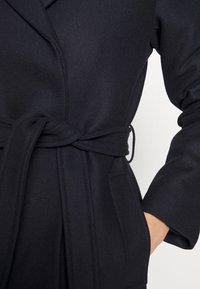 Gros plan d'une personne portant un manteau en laine noir noué à la taille avec une main dans la poche, sur un fond uni.