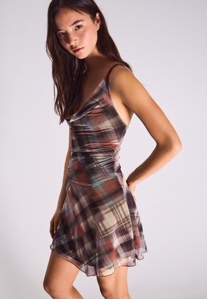 Kobieta z długimi ciemnymi włosami, ubrana w bezrękawnikową, wielokolorową sukienkę w kratę, stojąca z jedną ręką na biodrze na jednolitym tle.