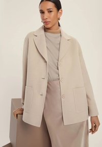 Cappotto in misto lana color beige con colletto a revers, bottoni frontali e due tasche; indossato su un top color taupe e una gonna in satin. Texture liscia.