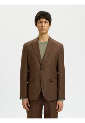 Jonge man met een bruine blazer met één knoop over een olijfgroene T-shirt met ronde hals, staand tegen een effen lichte achtergrond.