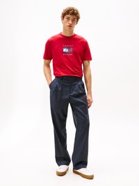Tricou roșu din bumbac cu grafică "Tommy Hilfiger", asortat cu pantaloni evazați de culoare albastră, dintr-un material texturat, și adidași albi cu tălpi maro.