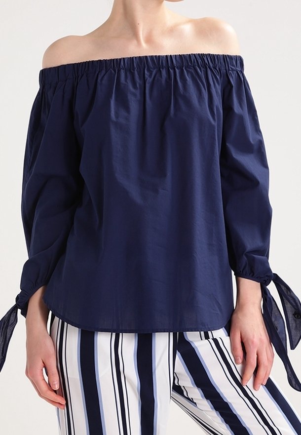 Haut bleu marine à épaules dénudées avec un col élastique, des manches longues et des accents de nœud aux poignets, associé à un pantalon taille haute rayé.