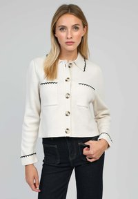 Veste en tricot blanche avec bord noir, grands boutons et poches, présentant un col et une silhouette cintrée.