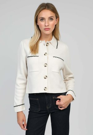 Veste en tricot blanche avec bord noir, grands boutons et poches, présentant un col et une silhouette cintrée.