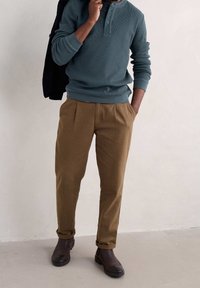 Pullover lavorato a maglia in teal con scollatura a girocollo e patta con bottoni, abbinato a pantaloni marroni a pieghe. Stivaletti neri alla caviglia completano l'outfit.