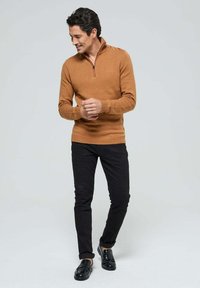 Pull en maille texturée, marron, avec fermeture zippée, associé à un pantalon noir et des mocassins noirs. Le modèle se tient debout avec les mains jointes.