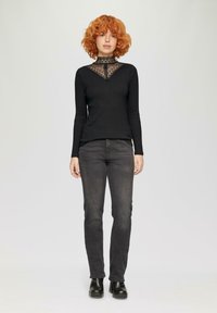 Schwarzes Langarmoberteil mit Spitzenverzierungen am Ausschnitt, kombiniert mit dunkelblauen Jeans und Plateaustiefeln, vor einem schlichten hellen Hintergrund.