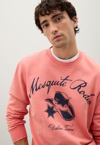 Sudadera coral con el texto "Mesquite Rodeo", gráfica de un sombrero vaquero y un diseño de estrellas; puños y cuello acanalados para añadir textura.