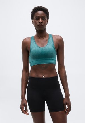 Persoană sportivă purtând bustieră sport teal și pantaloni scurți negri, stând în picioare pe un fundal simplu, deschis la culoare.