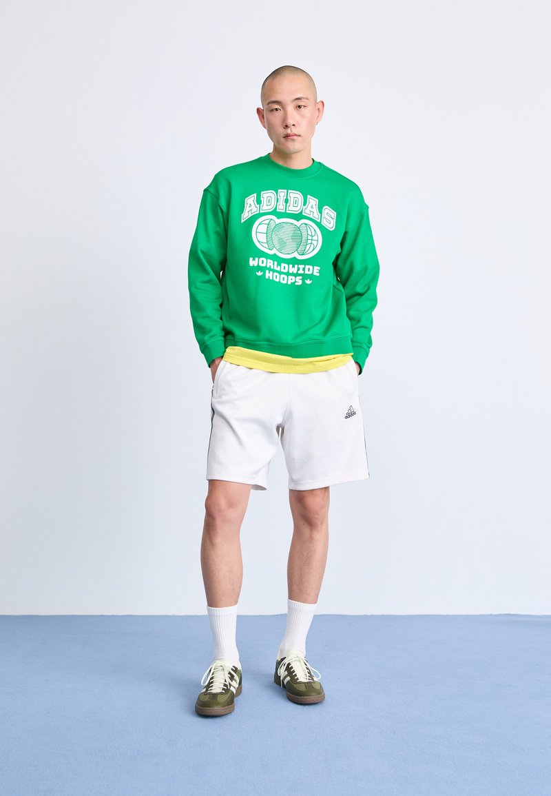 Groene sweatshirt met witte "ADIDAS" logo, gele onderhemd, witte shorts en groen/bruine sneakers. Eenvoudig ontwerp en relaxte pasvorm.