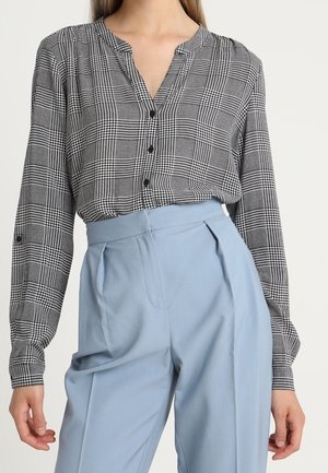 Femme portant un chemisier à carreaux noir et blanc rentré dans un pantalon plissé taille haute bleu clair.