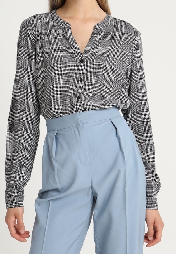 Femme portant un chemisier à carreaux noir et blanc rentré dans un pantalon plissé taille haute bleu clair.