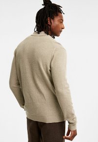 Beige stickad tröja med ribbade muddar och nederkant, med ett texturerat mönster och en klassisk krage. Sedd bakifrån, minimalistisk design.