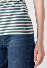 Dryžuotas t-shirt mėtų ir baltos spalvos, liesas kirpimas su trumpomis rankovėmis. Yra mažas logotipo akcentas šalia apvado, derinamas su mėlynos denim džinsais.