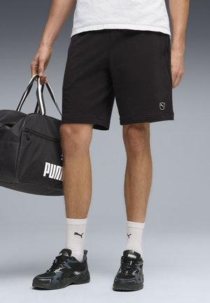 Uomo che indossa pantaloncini neri Puma, calzini bianchi Puma, scarpe da ginnastica nere Puma, che tiene una grande borsa da viaggio nera Puma per i manici.