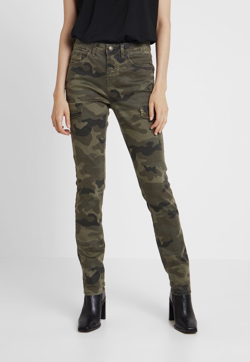 Personne portant un pantalon cargo camouflage slim, un haut sans manches noir et des bottines à talons noires, debout devant un fond uni.