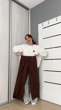 Pantaloni largi, de culoare maronie, cu talie elastică, susținuți de o persoană care poartă un hanorac alb. Textură moale, design lejer. Podea din lemn deschis.