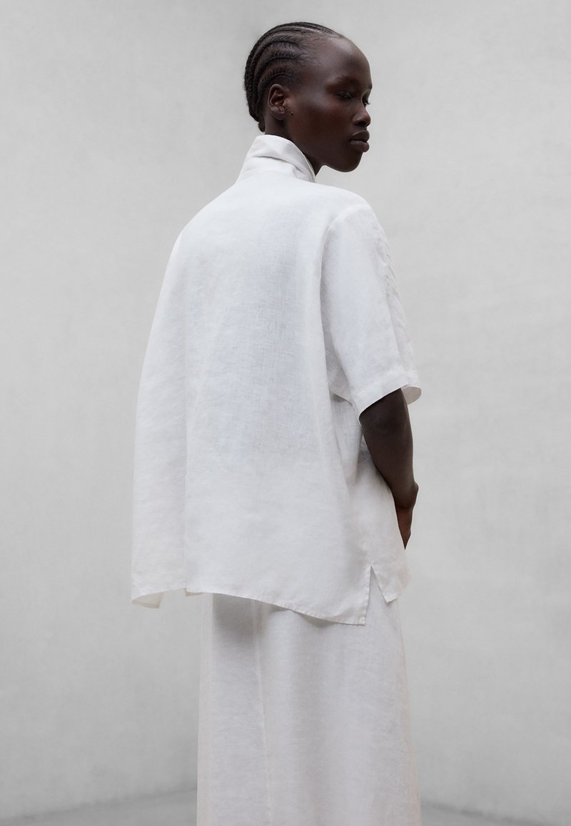 Femme aux cheveux tressés portant une chemise ample en lin blanc à manches courtes et un pantalon large assorti, sur un fond gris uni.