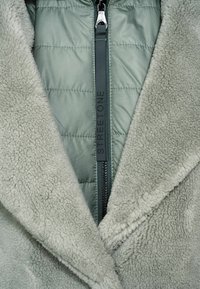 Ljusgrön fuskpälsjacka med dragkedja fram, quiltad textur på ärmarna och en metallic dragkedja med "STREETONE"-logotyp på dragfliken.